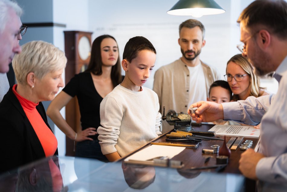 Ein Museumsmitarbeiter führt einer kleinen Gruppe Erwachsener und Kinder die traditionelle Uhrmacherei an der Werkbank eines Uhrmachers vor.