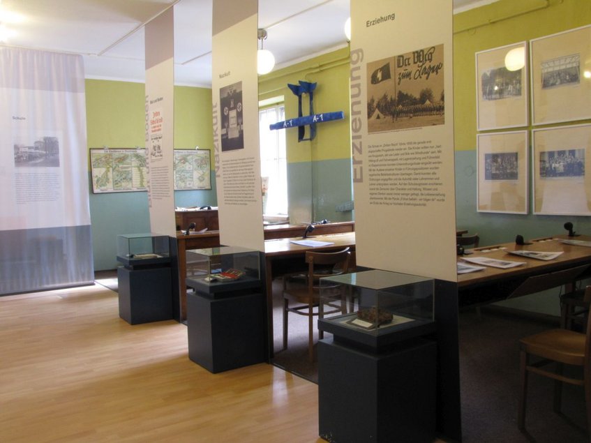 Foto eines Ausstellungsraums mit drei kleinen Tischvitrinen, drei Tischen, auf denen Informationsmaterial liegt, sowie mehreren Infotafeln beziehungsweise -bannern