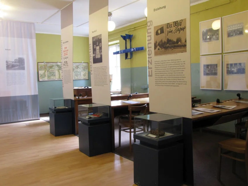 Ausstellung „Schule unterm Hakenkreuz“ Foto eines Ausstellungsraums mit drei kleinen Tischvitrinen, drei Tischen, auf denen Informationsmaterial liegt, sowie mehreren Infotafeln beziehungsweise -bannern