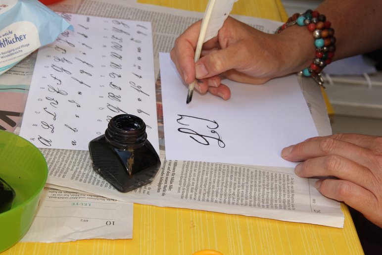 Das Foto zeigt die Hand einer weiblichen Person, die mit Gänsefeder und Tinte Buchstaben auf ein Blatt Papier schreibt.