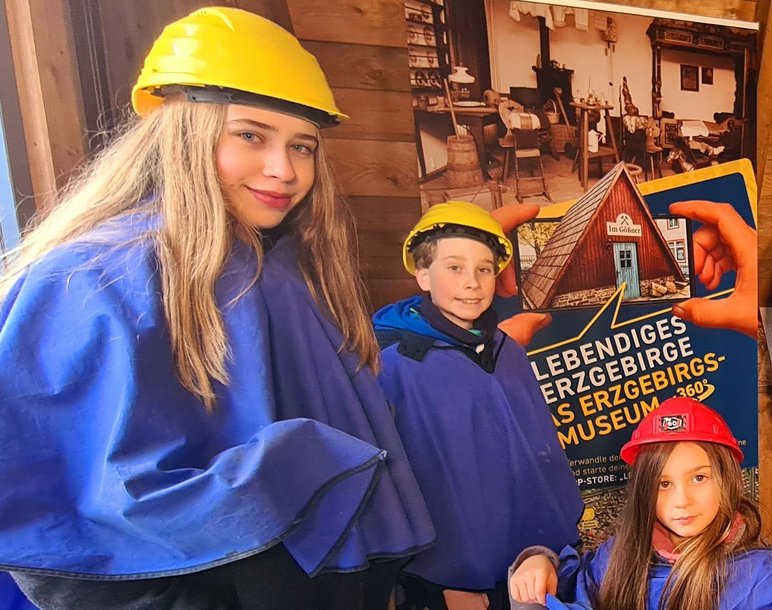 Das Foto zeigt drei Kinder im Besucherbergwerk „Im Gößner“, sie tragen gelbe Helme und dunkelblaue Umhänge.