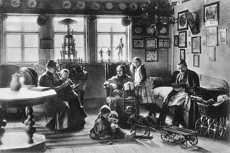 Historische Schwarzweißfotografie einer Wohnstube mit erzgebirgischer Volkskunst, ca. Anfang 20. Jahrhundert. In der Stube sitzt eine Familie zusammen, mittig im Bild sitzt eine alte Frau in einem Sessel, vor sich hat sie einen Klöppelsack.