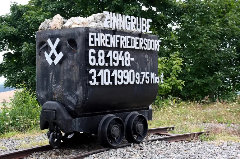Ein schwarzer Hunt (Kasten mit Rädern) auf Schienen mit Schotter vor einigen Bäumen. Auf dem Hunt ist an der vorderen kurzen Seite ein Zeichen (gekreuzt Eisen und Schlägel, auf dem Kopf stehend) und auf der langen vorderen Seite Schrift angebracht: "Zinngrube Ehrenfriedersdorf. 6.8.1948 - 3.10.1990 9.75 Mio t". Zeichen und Schrift sind weiß gehalten. Im Hunt liegen einige Felsbrocken.
