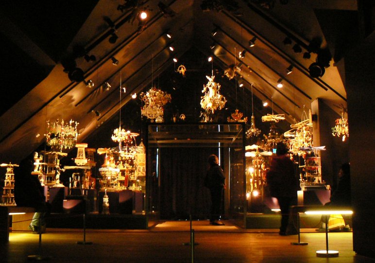 Das Foto zeigt zwei Menschen in einem beleuchteten Dachgeschossraum der Manufaktur der Träume. In dem Ausstellungsraum sind große handgeschnitzte Erzgebirgspyramiden zu sehen, und vom Spitzdach hängen handgeschnitzte Kronleuchter.