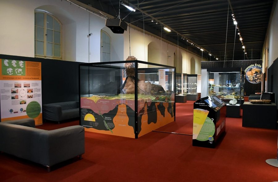 Das Foto zeigt einen Blick in die Ausstellung „Bewegte Erde“ mit einer Medienstation im Vordergrund sowie einer Vielzahl von Ausstellungsvitrinen und -tafeln.