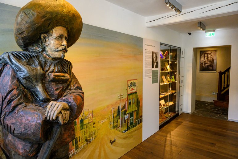 Das Foto zeigt den Eingangsbereich der Dauerausstellung mit einer lebensgroßen, aus Holz geschnitzten Figur von Karl May vor dem Wandbild einer Western-Szene. Rechts daneben schließen sich ein Ausstellungstext und eine beleuchtete Wandvitrine mit Büchern und Publikationen an.