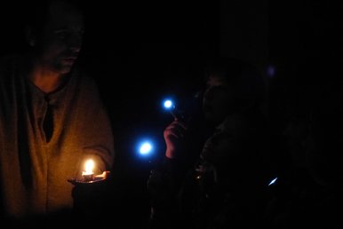 Ein erwachsener Mann mit Kerze und zwei Kinder mit Taschenlampen stehen, kaum erkennbar, in einem sehr dunklen Raum.