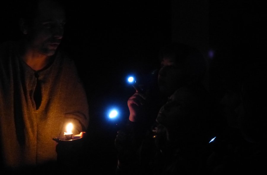 Ein erwachsener Mann mit Kerze und zwei Kinder mit Taschenlampen stehen, kaum erkennbar, in einem sehr dunklen Raum.