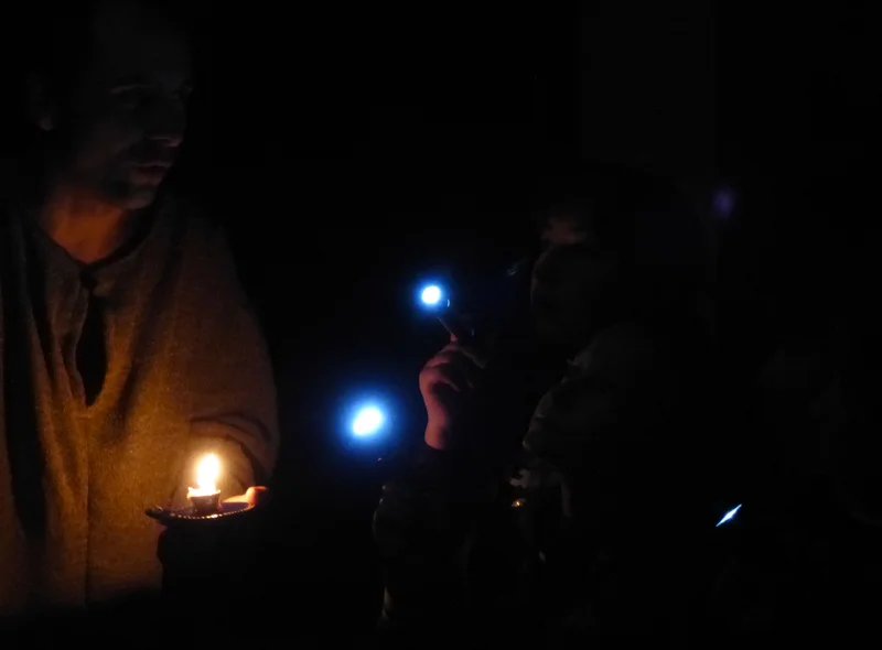 In einem dunklen Raum leuchten zwei Kinder mit kleinen Taschenlampen einen als Knecht verkleideten Mann mit Kerze an.