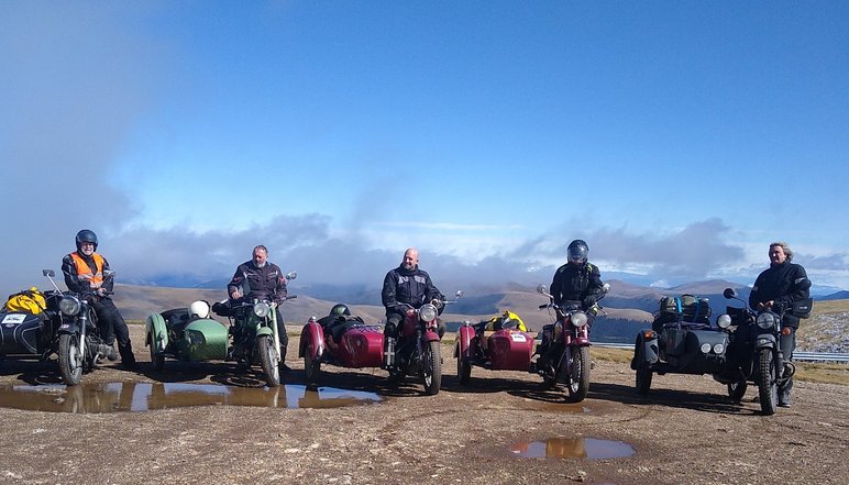 Fünf Motorradfahrer stehen nebeneinander mit ihren historischen Motorrädern mit Beiwagen in einer kargen Hochgebirgslandschaft.