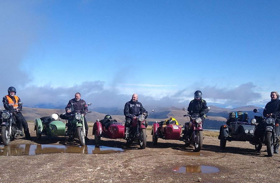 Fünf Motorradfahrer stehen nebeneinander mit ihren historischen Motorrädern mit Beiwagen in einer kargen Hochgebirgslandschaft.