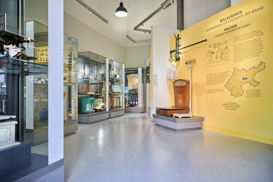 Foto des Eingangsbereichs des Waagenmuseums. Auf der großen, gelben Willkommenswand befindet sich neben diversen Texten eine Karte der Oschatzer Region, davor stehen zwei historische Personen- beziehungsweise Produktwaagen. Linkerhand reihen sich drei große Vitrinen mit einer Vielzahl historischer Waagen aneinander.