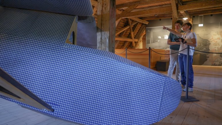 Zwei Personen auf einem historischen Dachboden mit hölzernem Gebälk, im Vordergrund ein großes Ausstellungsobjekt, das einen Segelflieger erahnen lässt