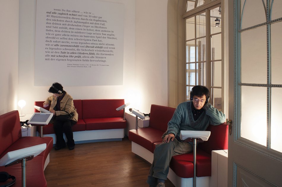 Das Foto zeigt zwei Besucher mit Kopfhörern, die, auf Bänken sitzend und auf Medienstationen blickend, im Hörkabinett des Bach-Museums Leipzig Stücken des bekannten Komponisten lauschen.