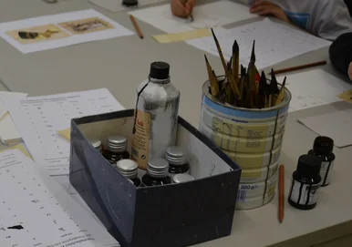 Auf einem Tisch liegen Schreibmaterialien: eine Schachtel mit Tintenfläschchen, ein Behälter mit verschiedenen Schreibfedern, ein Tintenfass sowie Blätter mit Übungsalphabeten und Schreibübungen.