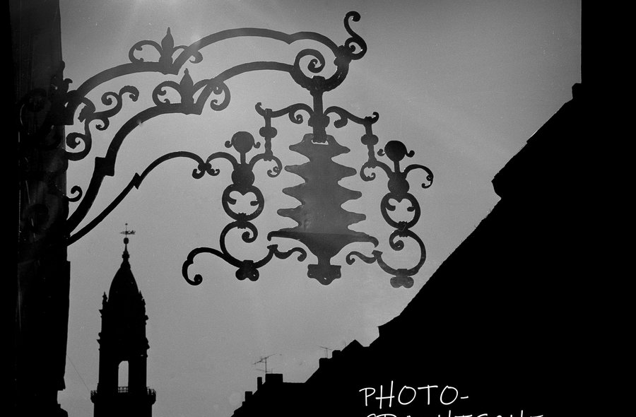 Gegen die Sonne und entlang einer Gebäudeseite in Richtung eines Kirchturmes fotografierte Schwarzweißaufnahme, in deren Vordergrund die Silhouette des Baumkuchenschildes eines Ladens oder Cafés abgelichtet ist
