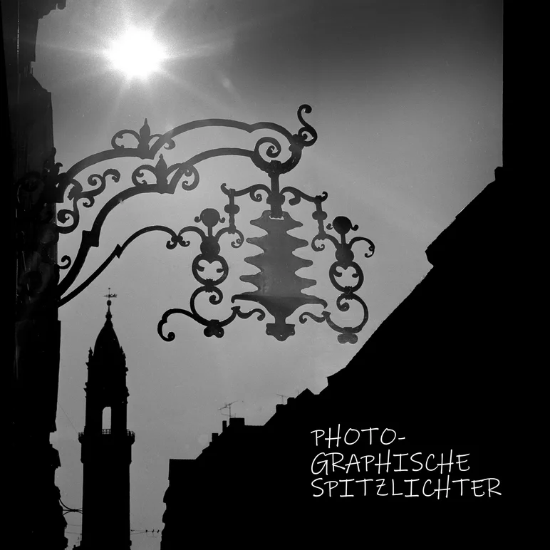 Gegen die Sonne und entlang einer Gebäudeseite in Richtung eines Kirchturmes fotografierte Schwarzweißaufnahme, in deren Vordergrund die Silhouette des Baumkuchenschildes eines Ladens oder Cafés abgelichtet ist