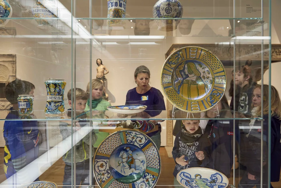 Eine Frau steht in einem Ausstellungsraum mit Vitrinen. Um sie herum sind viele Kinder, die ihr zuhören.