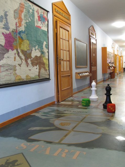 Foto eines langgestreckten Gangs im Schulmuseum, von dem mehrere Klassenzimmer abgehen; auf dem Boden stehen zwei überdimensionale Schachfiguren sowie zwei überdimensionale Würfel; an der Wand hängt eine historische Landkarte Europas.