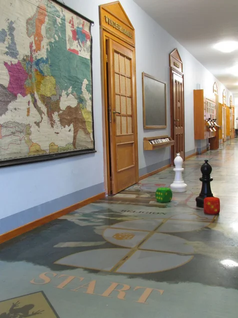 Fußbodenspiel in der „Kaiserzeitetage“ Foto eines langgestreckten Gangs im Schulmuseum, von dem mehrere Klassenzimmer abgehen; auf dem Boden stehen zwei überdimensionale Schachfiguren sowie zwei überdimensionale Würfel; an der Wand hängt eine historische Landkarte Europas.