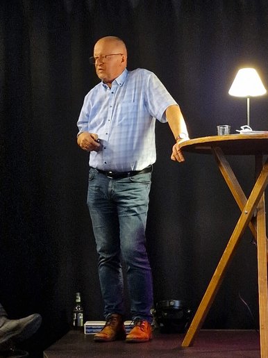 Ein Mann steht auf einer Bühne, an einen Stehtisch gelehnt, und wendet sich in Richtung Publikum.