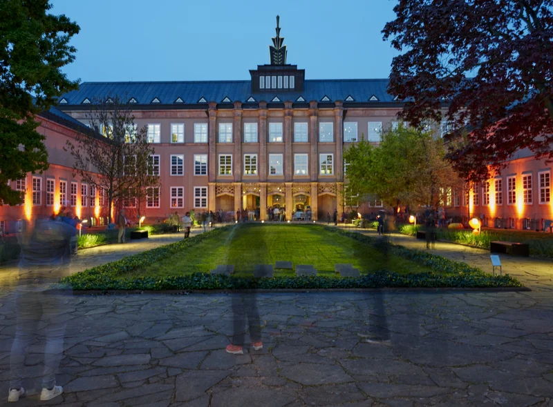 Museumsbau von außen zur blauen Stunde, mit Dachbekrönung. Im Vordergrund verschwommene Menschen, die ins Museum gehen.