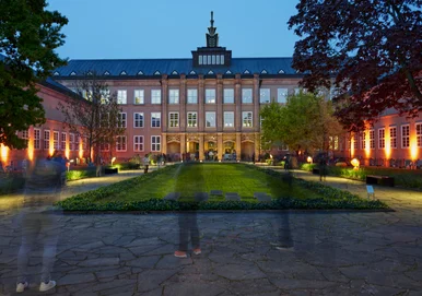 Museumsbau von außen zur blauen Stunde, mit Dachbekrönung. Im Vordergrund verschwommene Menschen, die ins Museum gehen.