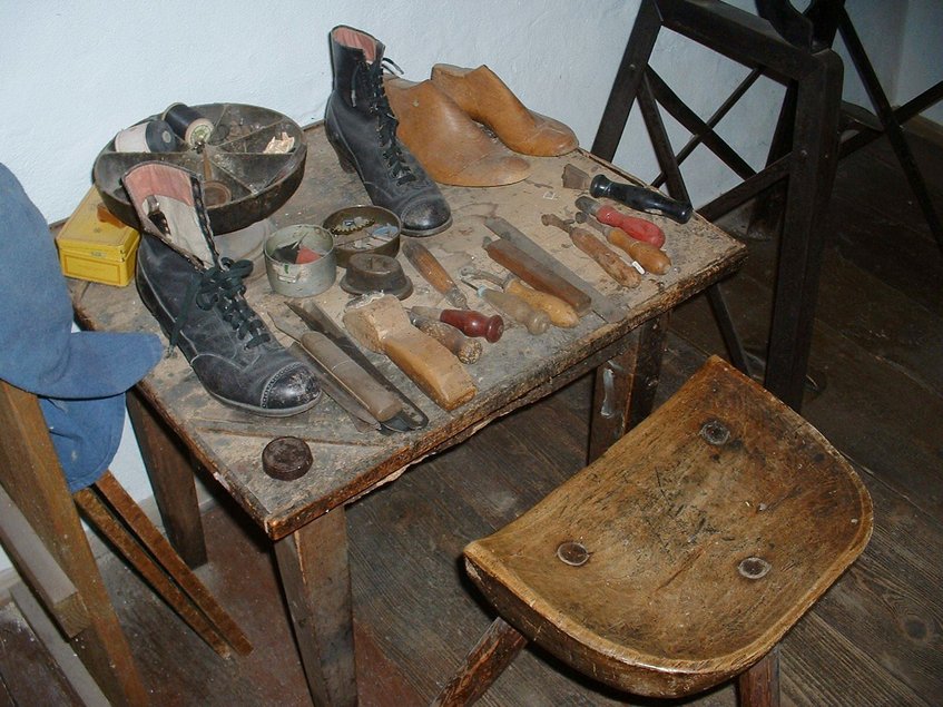 Historisch anmutender Arbeitsplatz des Schuhmachers: Auf einem einfachen, kleinen Holztisch befinden sich neben zwei Paar alter Schuhe diverse historische Schuhmacher-Werkzeuge. Vor dem Tisch steht ein hölzerner Schemel.
