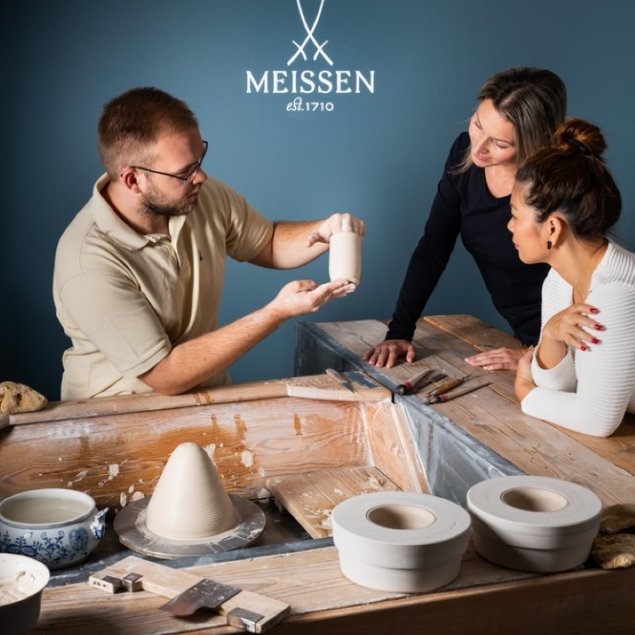 Ein Porzellanmacher zeigt zwei Frauen an einem Beispiel, wie der Vorgang der Porzellanherstellung erfolgt.