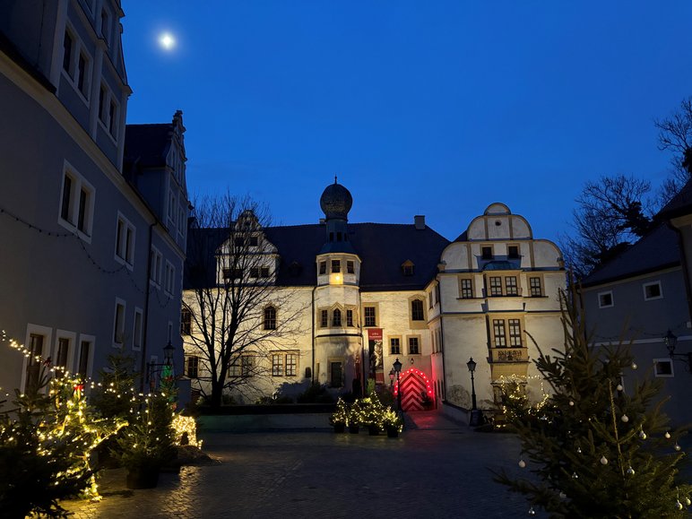 Spätabendliche Aufnahme eines historischen, weihnachtlich beleuchteten Schlosshofes. Am klaren, dunkelblauen Himmel scheint der Mond.