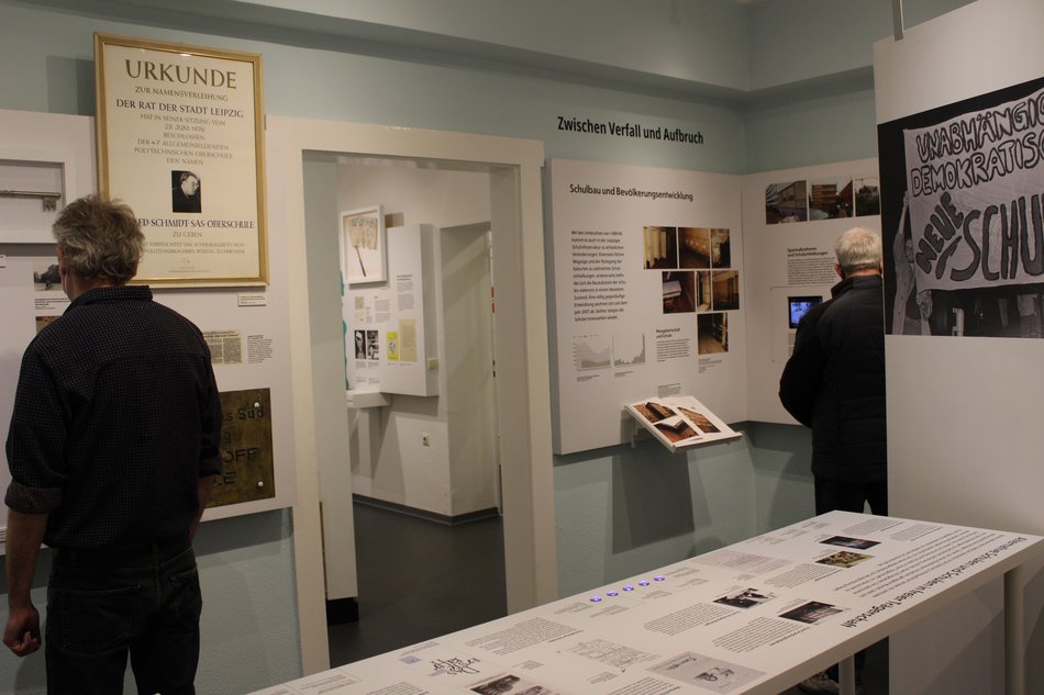 Foto eines Bereichs der Ausstellung zur Schule in Sachsen seit der politischen Wende von 1989/90. Zu sehen sind zwei erwachsene Besucher, die verschiedene Wandtexte studieren.