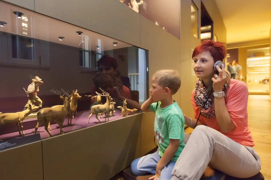 Eine Frau und ein Kind sitzen vor einer Vitrine mit einer Audiostation und hören eine Geschichte an.