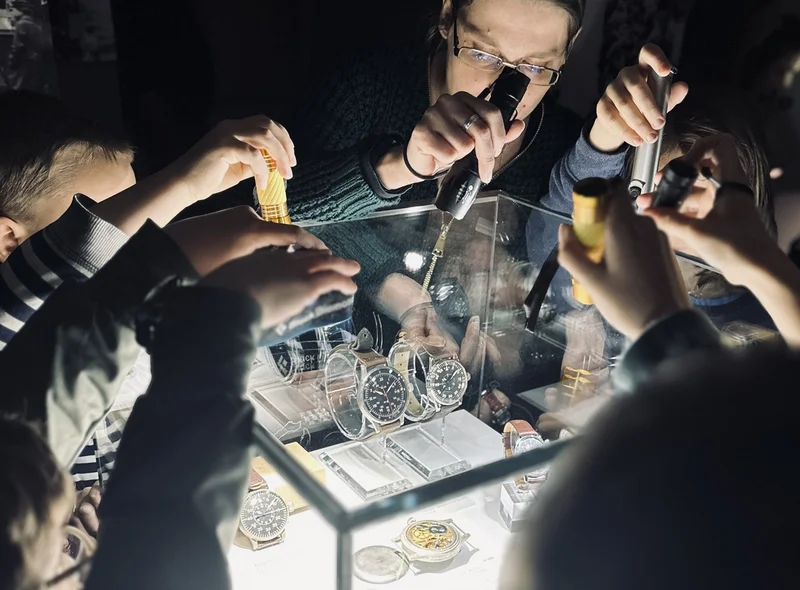 Kinder leuchten mit Taschenlampen von oben in eine Vitrine, in der Taschenuhren und Armbanduhren präsentiert werden.