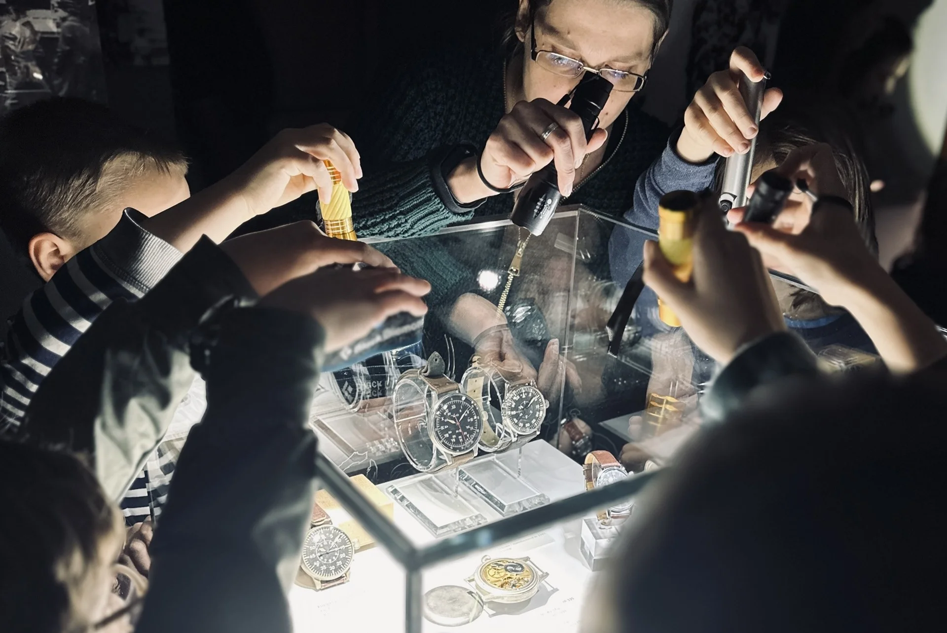 Kinder leuchten mit Taschenlampen von oben in eine Vitrine, in der Taschenuhren und Armbanduhren präsentiert werden.