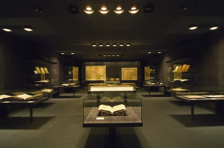 Zu sehen ist die Schatzkammer aus der Perspektive der Eintrittstür. Der Raum ist relativ dunkel. Links, mittig und rechts befinden sich Vitrinen, in denen alte Bücher zu erkennen sind. Hinten befindet sich eine große Wandvitrine, in der große, alte Landkarten ausgestellt sind.