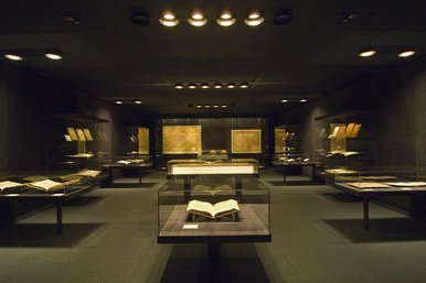 Blick in die Schatzkammer des Buchmuseums der SLUB Dresden Zu sehen ist die Schatzkammer aus der Perspektive der Eintrittstür. Der Raum ist relativ dunkel. Links, mittig und rechts befinden sich Vitrinen, in denen alte Bücher zu erkennen sind. Hinten befindet sich eine große Wandvitrine, in der große, alte Landkarten ausgestellt sind.
