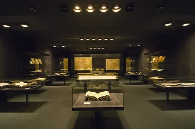 Zu sehen ist die Schatzkammer aus der Perspektive der Eintrittstür. Der Raum ist relativ dunkel. Links, mittig und rechts befinden sich Vitrinen, in denen alte Bücher zu erkennen sind. Hinten befindet sich eine große Wandvitrine, in der große, alte Landkarten ausgestellt sind.