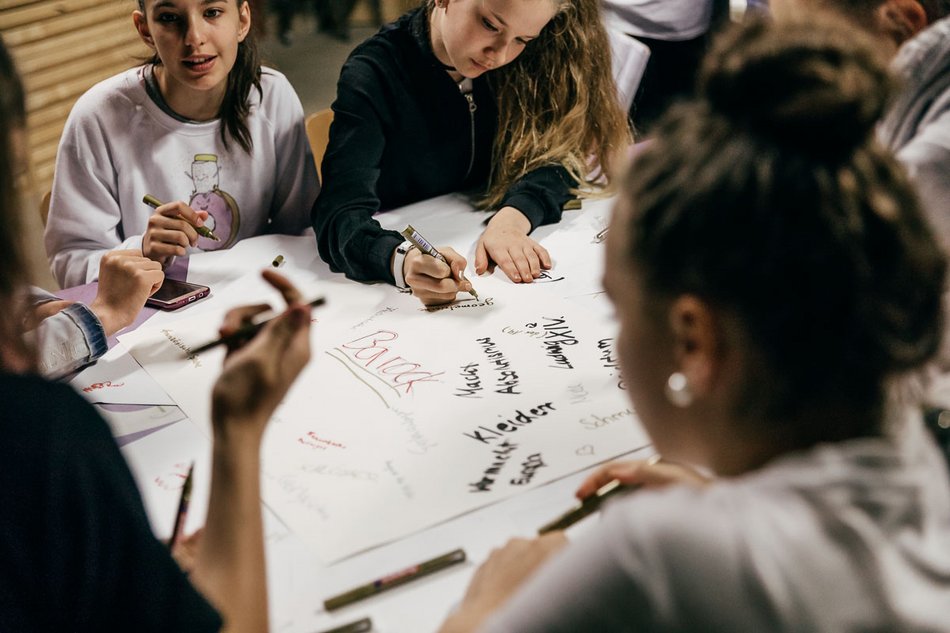 Das Foto zeigt die Stifte haltenden Hände mehrerer Jugendlicher über einem weißen Papierbogen, auf dem in schwarzer und weinroter Farbe Worte wie „Barock“, „Kleider“, „Macht“ und „Absolutismus“ geschrieben wurden.