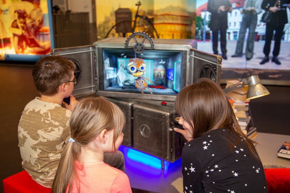 Von hinten sind drei Kinder zu sehen, die vor einer mit einem Bildschirm ausgestatteten, in spielerischem Industrie-Look gestalteten Medienstation sitzen. Auf dem Bildschirm ist die Figur eines kleinen Roboters erkennbar.