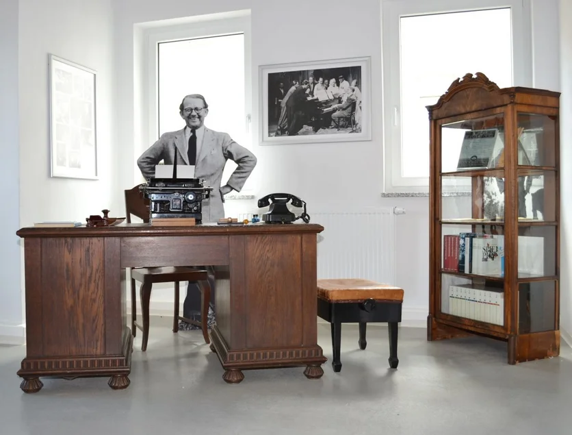 In einem weiß getünchten Ausstellungsraum steht hinter einem großen historischen Holzschreibtisch die lebensgroße Figur eines älteren, lächelnden Mannes. Auf dem Schreibtisch befinden sich eine alte Schreibmaschine und ein historisches Telefon. Am rechten Bildrand ist eine historische Vitrine mit Büchern und Bildern zu sehen.