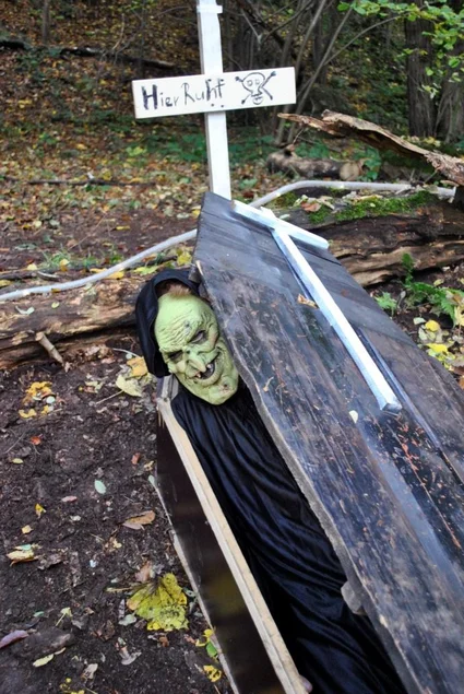 Aus einer geöffneten Sarg-Attrappe lugt die mannsgroße Puppe eines Toten. Auf dem Grabstein dahinter steht "Hier ruht…"