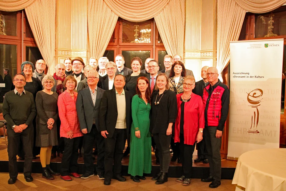 Foto einer drei-reihig angeordneten Gruppe von Frauen und Männern in einem Festsaal; am rechten Bildrand ist ein Banner mit der Aufschrift "Ehrenamt in der Kultur" zu sehen.