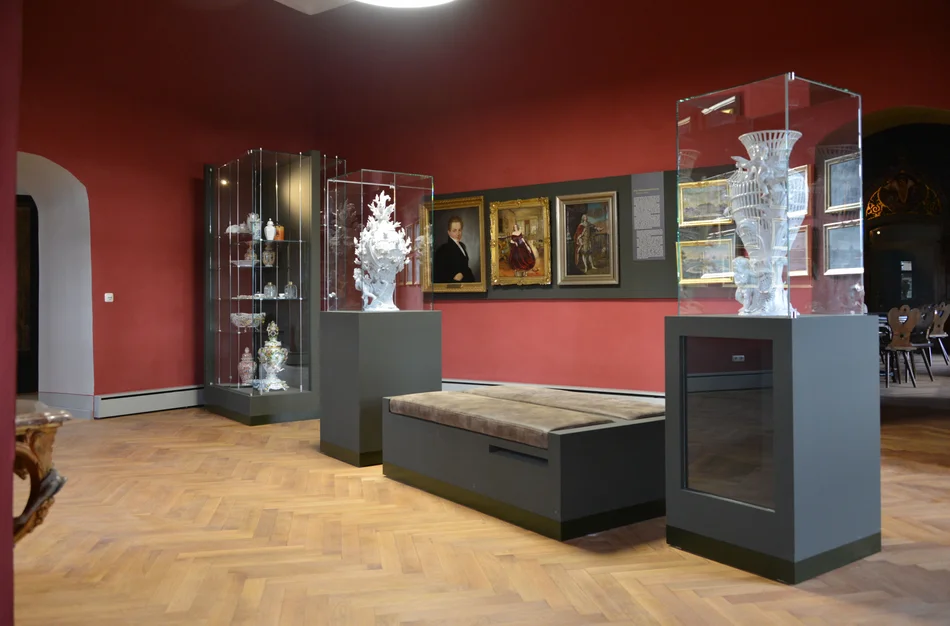 Drei Vitrinen mit weißen Porzellanvasen, an der Wand dahinter hängen verschiedene Bilder.