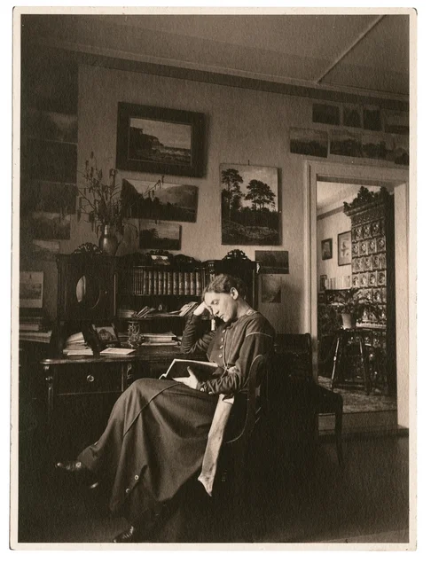 Fotografie einer lesenden Frau vor einem Schreibtisch mit Schrank-Aufsatz sitzend. Ihr Kopf ist auf den rechten Arm gestützt.