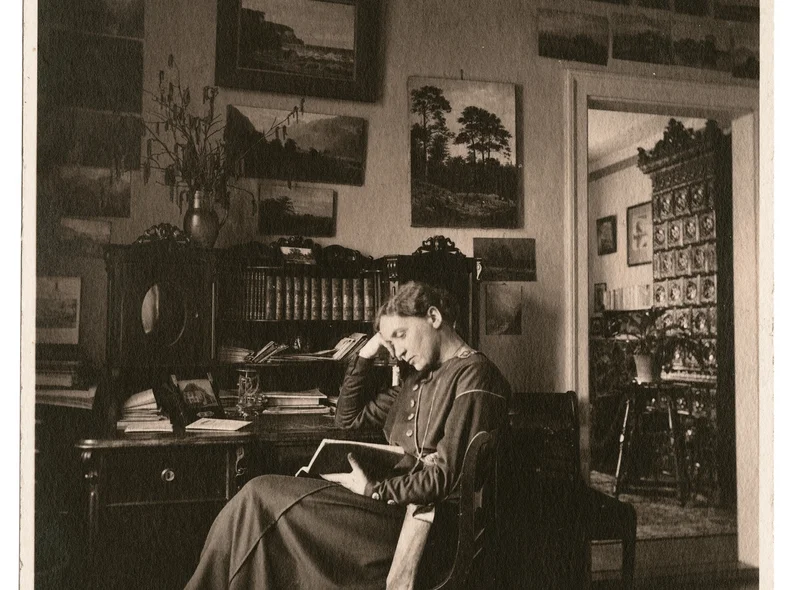 Fotografie einer lesenden Frau vor einem Schreibtisch mit Schrank-Aufsatz sitzend. Ihr Kopf ist auf den rechten Arm gestützt.