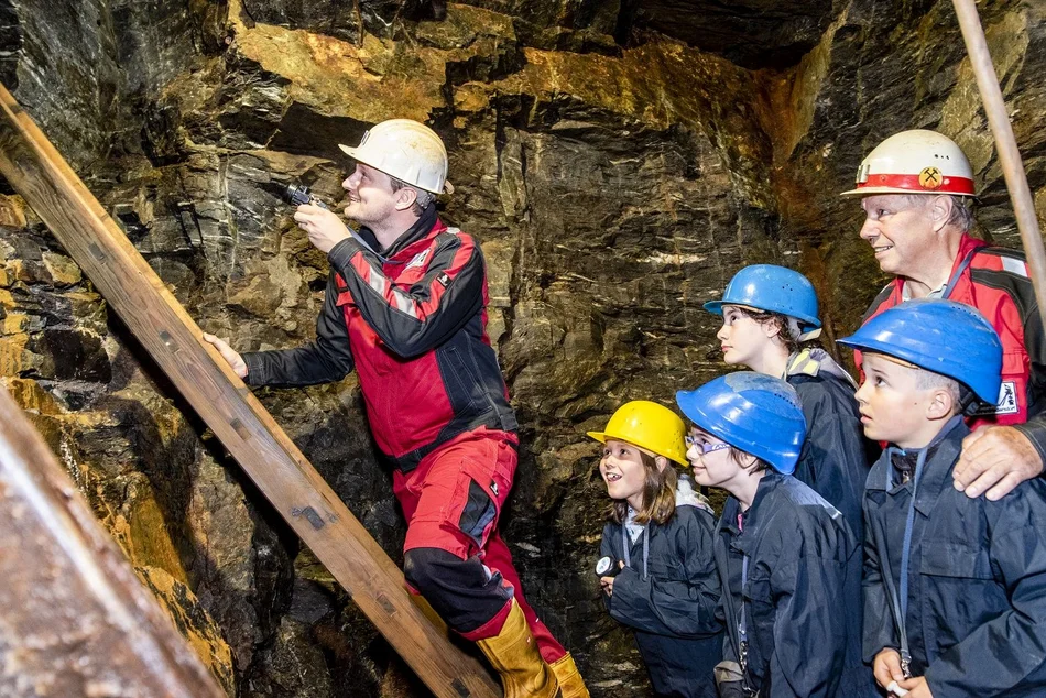 Bergbau erleben für Groß und Klein Zwei Männer und vier Kinder in Schutzkleidung und mit Helmen im Besucherbergwerk, einer der Männer scheint mit einer Taschenlampe den Weg eine Leiter hinauf.