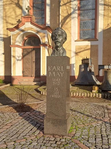 Vor einer Kirche steht die Büste eines bärtigen Mannes mit der Sockel-Inschrift "Karl May. 1842 bis 1912. Es sei Friede".