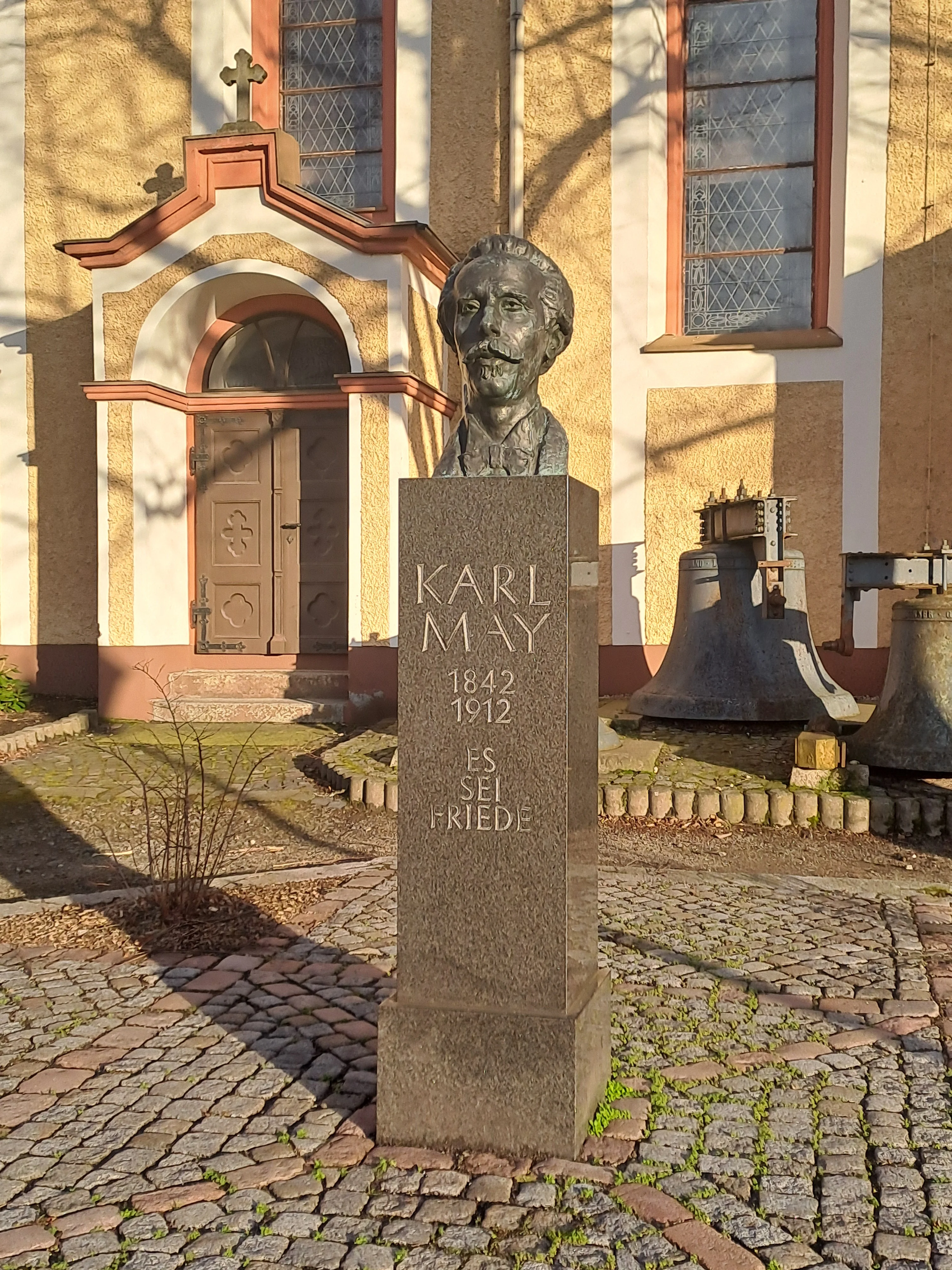 Vor einer Kirche steht die Büste eines bärtigen Mannes mit der Sockel-Inschrift "Karl May. 1842 bis 1912. Es sei Friede".
