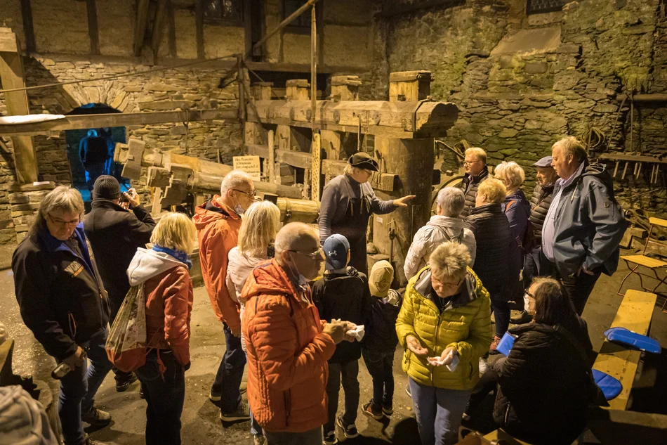 Eine Besuchergruppe und ein Museumsführer stehen im Inneren eines alten Natursteingebäudes, im Hintergrund sind große historische Schmiedehämmer erkennbar.