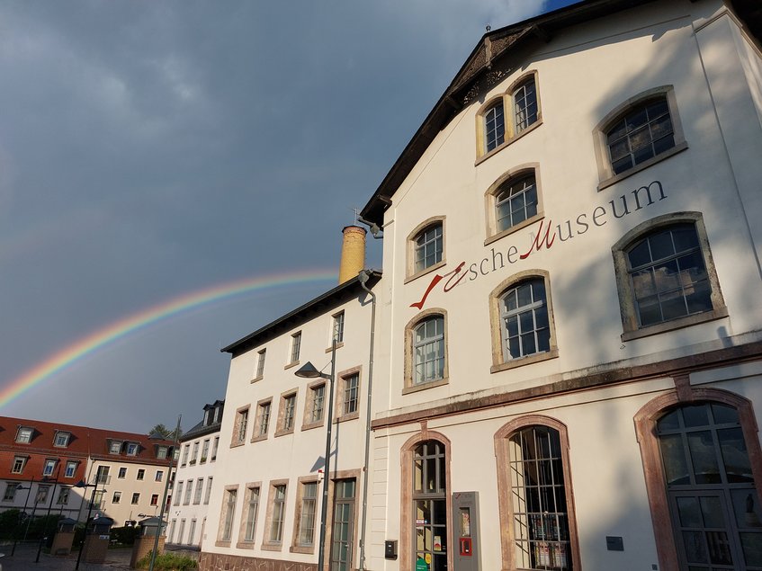 Das Foto zeigt eine Außenansicht des Esche-Museums in Limbach-Oberfrohna, welches sich in einem im 19. Jahrhundert errichteten Gebäude der ehemaligen Wirkerei der Familie Esche befindet. Den düsteren Himmel im Hintergrund überspannt ein Regenbogen.